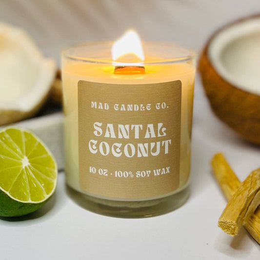 Scented candle labeled 'Santal Coconut' with coconut halves, a lime, and a stick of cinnamon on a light background.